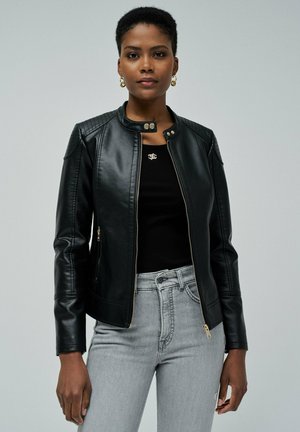 Veste en cuir noire avec un design ajusté, col rond, fermeture éclair et accents matelassés aux épaules. Portée sur un haut noir et un jean gris.