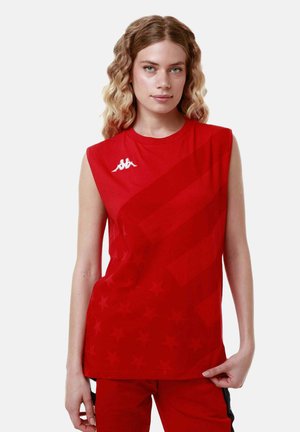 Giovane donna con capelli biondi ondulati che indossa un top rosso senza maniche con un motivo sottile a stelle e un logo bianco, in piedi su uno sfondo bianco.