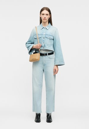 Femme portant une veste en jean bleu clair et un jean, tenant un sac bandoulière beige, avec une ceinture noire et des bottines noires, debout sur un fond blanc.