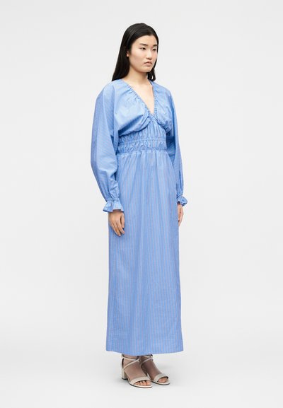 Γυναίκα στέκεται φορώντας ένα μακρύ μπλε ριγέ φόρεμα με βαθύ V-ντεκολτέ, σφηνοειδική μέση, μακριά φουσκωτά μανίκια και μπεζ ανοιχτές γόβες.