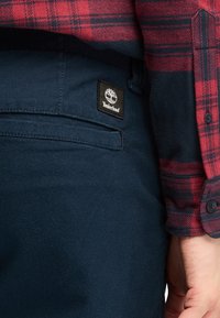 Navy blue pants with a smooth texture and a visible black Timberland logo patch. A red and black plaid shirt is partially visible above.