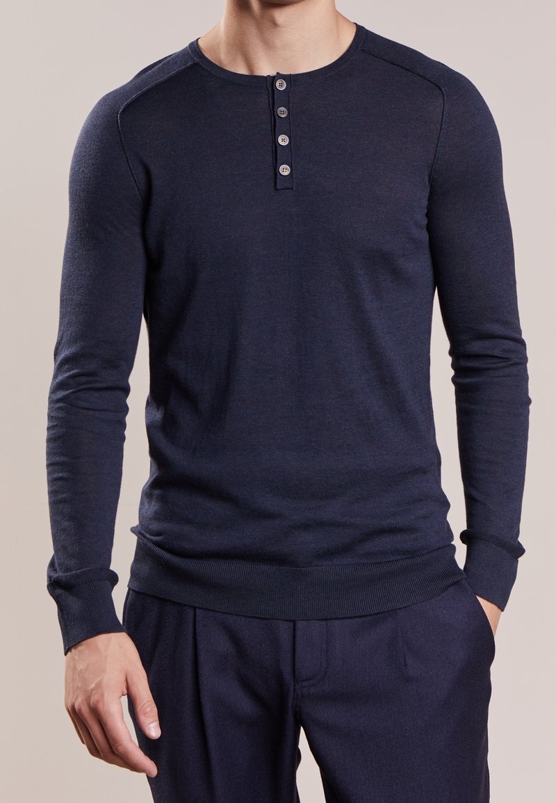 Homme portant un t-shirt henley ajusté manches longues bleu marine foncé avec quatre boutons et pantalon foncé assorti, mains détendues le long du corps.