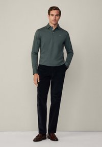 Dlouhý rukáv, zelené polo tričko s límečkem, spárované s černými kordovými kalhotami. Hnědé boty dotvářejí outfit.