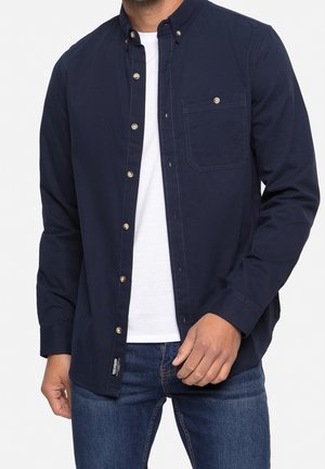 Mann trägt ein dunkelblaues Hemd mit Knöpfen über einem weißen T-Shirt und blauen Jeans, steht mit einer Hand nahe der Taille.