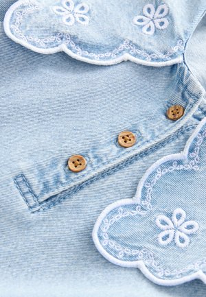 Lichtblauwe denimstof met geschulpte randen en witte borduursels van bloemmotieven, versierd met vier houten knopen langs de naad.