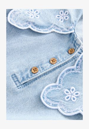 Tessuto di denim azzurro chiaro con bordi scallopati e ricami bianchi, decorato da motivi floreali e quattro bottoni in legno lungo la cucitura.