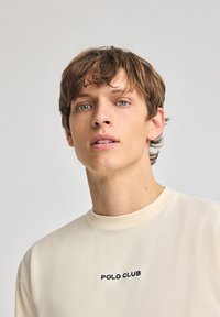Sudadera color crema con cuello redondo y puños acanalados. Presenta bordado en negro de "POLO CLUB" en el pecho. Textura suave, diseño casual.