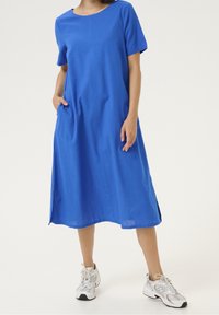 Sininen lyhythihainen mekko, valmistettu kevyestä kankaasta, jossa pyöreä kaula-aukko ja sivutaskut. Yhdistetty valkoisiin lenkkareihin.