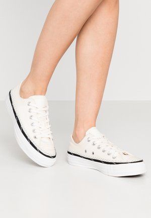 Close-up van de onderbenen van een persoon die witte lage sneakers met zwarte bies draagt, staand op een lichtgrijze vloer.