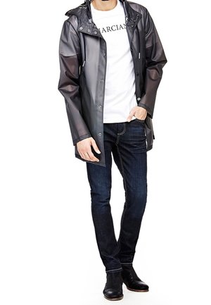 Impermeabile grigio con cappuccio, bottoni a pressione e tasche laterali, indossato sopra una maglietta bianca e jeans scuri. Stivali neri alla caviglia.