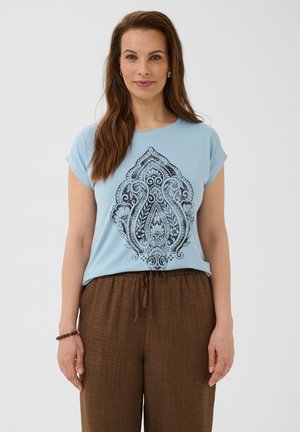 Femme aux longs cheveux bruns portant un t-shirt bleu avec un motif noir ornementé et un pantalon marron ample, debout devant un fond clair uni.