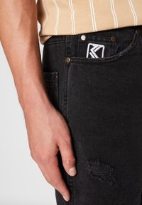 Mustat farkut, joissa on hieman kulutusta, sisältäen taskun, jossa on valkoinen logopatch ja kultaisia yksityiskohtia. Tekstuuri on sileä ja leikkaus on kapea.