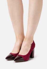 Chaussures à talons hauts bordeaux avec un corps en daim et des bouts pointus en cuir verni. Le talon carré offre stabilité et structure.