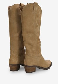 Botas de ante color beige hasta la rodilla con punta redondeada, tacón de madera apilado y costuras en contraste; cuentan con un tirador en la parte superior para facilitar su uso.