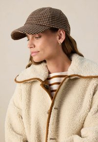 Bruin houndstooth pet met een gebogen klep, gecombineerd met een crèmekleurige schapenvachtjas met een tan kraag en knoopsluiting, gelaagd over een gestreept shirt.