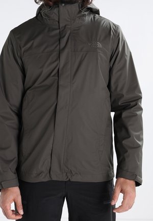 Uomo che indossa una giacca impermeabile verde scuro North Face con cappuccio e pantaloni neri, su sfondo neutro.