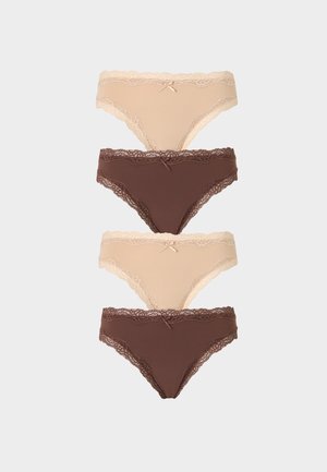 Femme aux longs cheveux bruns portant un soutien-gorge beige orné de dentelle et un slip assorti, debout devant un fond clair uni.