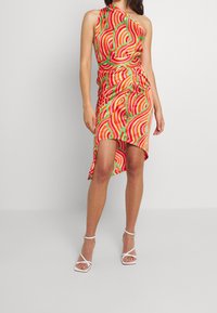 Robe colorée à une épaule avec des motifs tourbillonnants rouges, roses et verts. Tissu léger, ourlet asymétrique, détail de taille nouée, associée à des talons blancs.