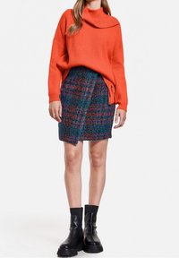 Pomarańczowy prążkowany sweter z szerokim kołnierzem, zestawiony z wielokolorową, kratkowaną spódnicą typu tweed. Czarne botki za kostkę z masywną podeszwą.