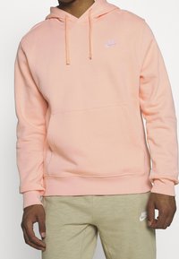 Man i en ljusrosa Nike-hoodie med framficka och beige Nike-mjukisbyxor, står med avspända armar.