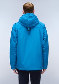 Uomo che indossa una giacca a capuccio blu brillante, rivolto di spalle contro uno sfondo grigio chiaro.