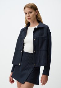 Giacca a righe navy in tessuto, con colletto, bottoni a pressione dorati e due tasche frontali. Abbinata a una gonna coordinata.
