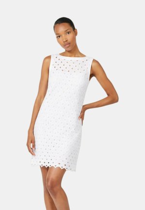 Donna che indossa un vestito bianco senza maniche con ritagli a forma di diamante, in piedi con una mano sul fianco su sfondo semplice.
