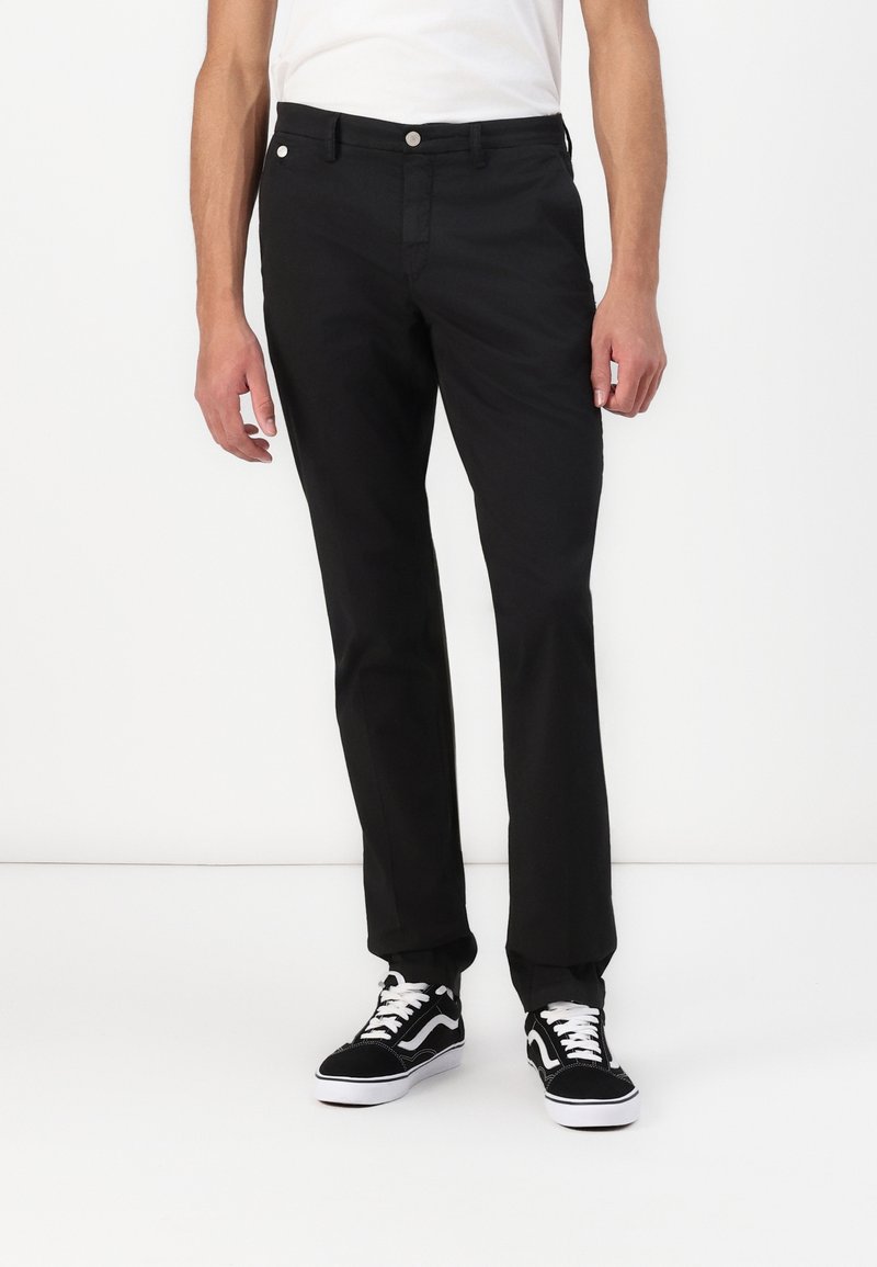 Pantalon noir ajusté avec une texture lisse, doté d'une fermeture par bouton et zip. Porté avec des baskets noires et blanches, présenté sur un fond uni.
