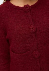 Bordeauxfärgad cardigan med strukturerad stickning, som har en rundad halsringning, tre blommformade knappar och en enkel frontficka.