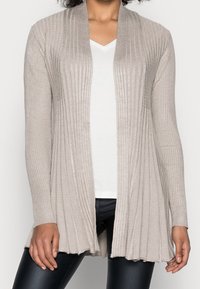 Cardigan beige a coste con apertura frontale, maniche lunghe e finitura testurizzata. Indossato sopra una t-shirt bianca a scollo a V e leggings neri.