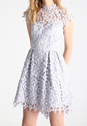 Biała, koronkowa sukienka bez rękawów z kwiatowym wzorem, z wysokim kołnierzem, dopasowaną talią i rozkloszowanym dołem sięgającym ponad kolano na stojącej osobie.