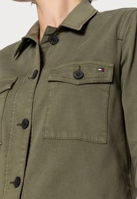 Jacket olivové barvy, vyrobený z hladkého materiálu. Obsahuje dvě klopové kapsy, černé knoflíky a malou tri-barevnou logo akcentaci.