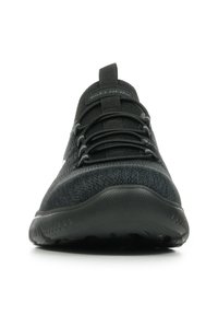 Zapatilla deportiva negra con parte superior de punto texturizado, suela flexible y cordones elásticos, con un diseño minimalista y cuello acolchado.