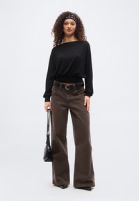 Νεαρή γυναίκα με σγουρά μαλλιά, φορώντας μαύρη κορδέλα, μαύρο μακρυμάνικο τοπ, καφέ παντελόνι φαρδιάς γραμμής, μαύρη ζώνη και κρατώντας μαύρη τσάντα χειρός.