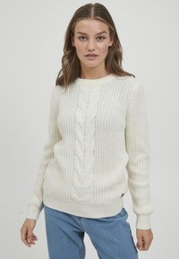 Hvit strikket genser med kabeldesign nedover midten, ribbestrikkede mansjetter og hem, og et strukturert mønster. Kombinert med blå jeans.