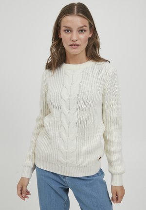 Hvit strikket genser med kabeldesign nedover midten, ribbestrikkede mansjetter og hem, og et strukturert mønster. Kombinert med blå jeans.