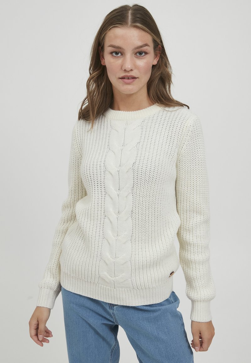 Hvit strikket genser med kabeldesign nedover midten, ribbestrikkede mansjetter og hem, og et strukturert mønster. Kombinert med blå jeans.