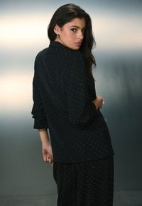 Čierna košeľa na zapínanie s jemnými bielymi bodkami, dlhými rukávmi a voľným strihom. Tkanina má hladkú textúru a mierny lesk.