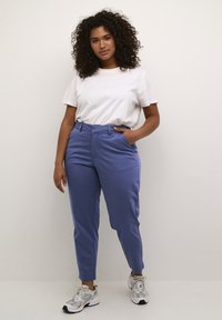 Pantaloni su misura blu chiaro con tasche laterali, indossati con una t-shirt bianca. Modello che indossa scarpe da ginnastica grigie e bianche, in posizione eretta contro uno sfondo neutro.