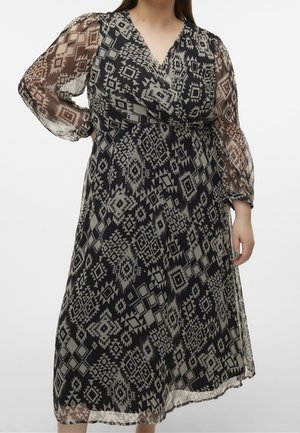 Kobieta ubrana w midi sukienkę w geometryczny wzór w kolorach czarnym i beżowym, z przezroczystymi długimi rękawami i kopertowym dekoltem w kształcie litery V.