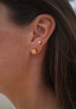 Nahaufnahme eines Frauenohres mit zwei quadratischen goldenen Ohrsteckern, einem kleineren über einem größeren, vor dunkelbraunem Haar.