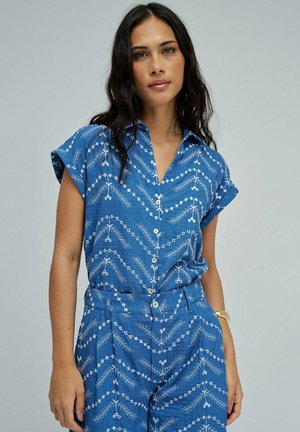 Mujer con cabello largo y oscuro, que lleva una camisa azul de manga corta con botones y pantalones de cintura alta a juego, ambos con patrones de hojas geométricas blancas.