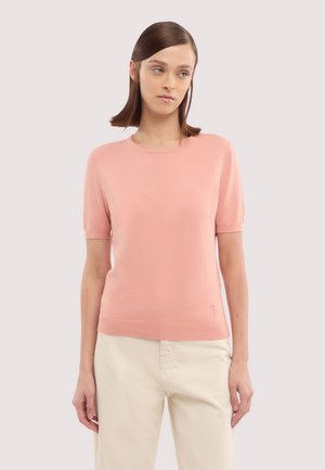 Ung kvinde med glat brunt hår iført en lyserød strikket bluse med korte ærmer og lyse beige bukser, der står mod en ensfarvet lys baggrund.