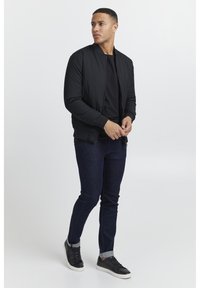 Giubbotto bomber nero realizzato in materiale leggero, con zip frontale, polsini a costine e un design elegante, indossato sopra una maglietta nera e jeans scuri.