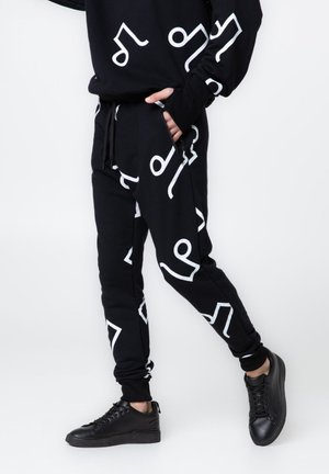 Osoba ubrana w czarne spodnie dresowe i bluzę z białymi abstrakcyjnymi kształtami, czarne trampki, ręka w kieszeni, na białym tle.