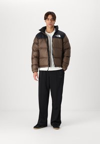 Jachetă puffer de culoare maronie cu umeri negri, închidere cu fermoar și logo. Asortată cu pantaloni negri și adidași. Textură netedă, design scurt.