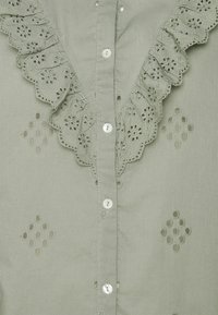 Lichtgroene blouse met een rimpelrand, geborduurd bloempatroon, decoratieve uitsparingen en parelachtige knopen aan de voorkant.