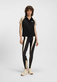Svart och beige kortärmad polo med knappar och logotyp, tillsammans med högmidjade svarta leggings med beiga och vita ränder och detaljer.