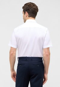 Camicia bianca a maniche corte con colletto standard, texture liscia, abbinata a pantaloni blu navy con tasche posteriori decorate da bottoni.