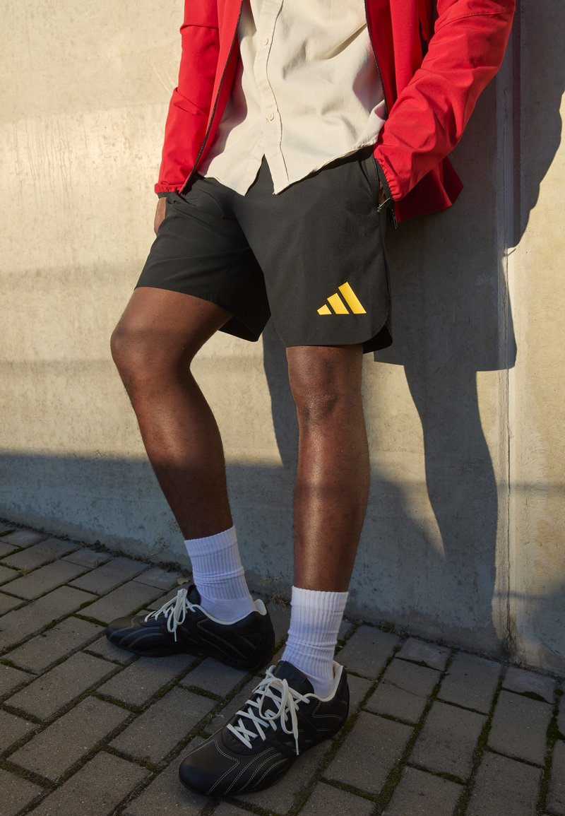 Melni sporta šorti ar dzelteno Adidas logo, valkāti ar gaišu krekliņu un sarkanu jaku. Melni sporta apavi ar baltām auklām un zeķēm.